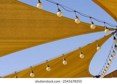 Awning texture of triangle fabric stretch roof made of canvas sail with rows of hanging bulb lamps for party night lighting on blue sky background. Outdoors lamp strings and nylon sun shades. - Powered by Shutterstock