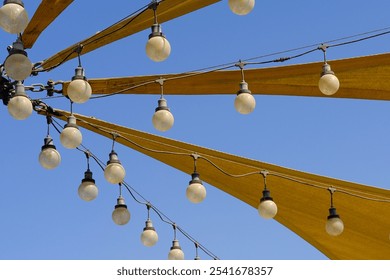 Awning texture of triangle fabric stretch roof made of canvas sail with rows of hanging bulb lamps for party night lighting on blue sky background. Outdoors lamp strings and nylon sun shades. - Powered by Shutterstock