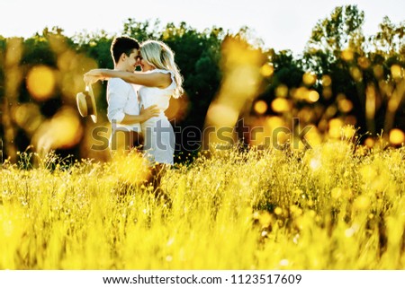 Awsome happy couple have fun in a field sunset background. Handsome Caucasian guy wearing white shirt gentle hugs a smiling girl with blond hair in white dress. Lifestyle and travel concept.