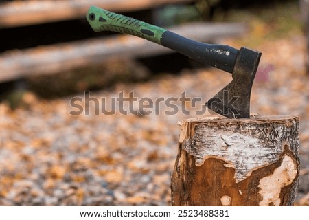 An axe will be stuck into a wooden log. Harvesting firewood for the winter season. Logging on the background of a country house. Lumber, wood-cutting tools.