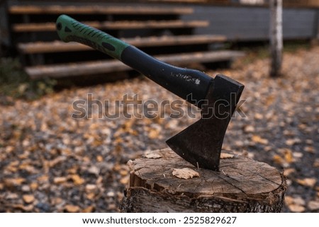 An axe will be stuck into a wooden log. Harvesting firewood for the winter season. Logging on the background of a country house. Lumber, wood-cutting tools.