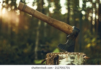 An axe with a wooden handle is stuck in a wooden log. Logging on the background of sunset. The hatchet.
