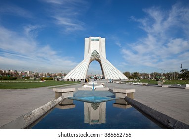 Azadi monument and its reflection on waterways in Azadi square of Tehran, against blue sky and white clouds.
