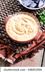 An Aztec Bowl Of Vegan Spicy Cheese Dip On Textured, Woven Place Mat With Bowl Of Blue Corn Tortilla Chips And Fresh Cilantro In The Backgorund.