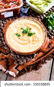 An Aztec Bowl Of Vegan Spicy Cheese Dip On Textured, Woven Place Mat With Bowl Of Blue Corn Tortilla Chips And Fresh Cilantro In The Background.