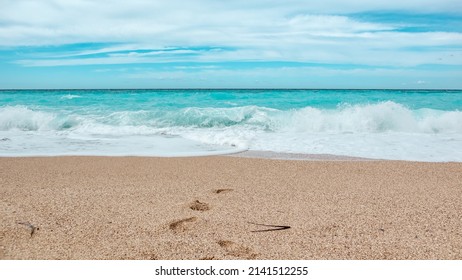 Azure Colored Waves With Blue Sky On Sunny Coast Of Greek Island. Sandy Beach With Footprints In Greece. Summer Nature Vacation Travel To Ionian Sea