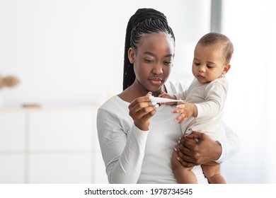 Babies And Fever. Young Black Mom Measuring Toddler's Temperature With Thermometer Caring For Ill Son, Holding Kid On Hands Standing In Bedroom At Home. Child Care And Infant's Health Problem Concept