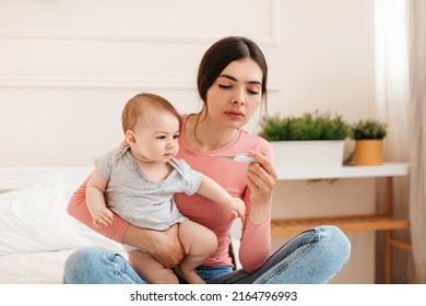 Babies and fever. Young mother measuring kid's temperature with thermometer, caring for ill daughter, sitting on bed in bedroom at home. Child care and infant's health problem concept