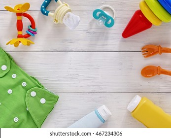 Babies Goods: Cloth Diaper, Baby Powder, Nibbler, Cream, Teether, Soother, Baby Spoon And Fork, Pyramid Toy On White Wooden Background.copy Space.Top View. Early Childhood Development Concept