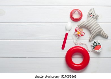 Baby Accessories Background: Socks, Soother, Nipple, Spoon And Toys Over White Wooden Background With Copy Space; Top View, Flat Lay
