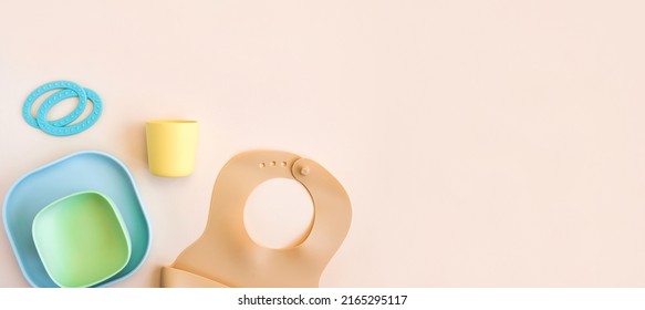 Baby Accessories And Tableware For Food On Beige Background. First Feeding For Kids Concept. Flat Lay, Top View, Space For Text. Silicone Dishware, Bib, Cup On Neutral Backdrop. Serving Baby Food.