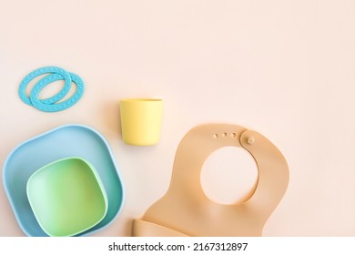 Baby Accessories And Tableware For Food On Beige Background. First Feeding For Kids Concept. Flat Lay, Top View, Space For Text. Silicone Dishware, Bib, Cup On Neutral Backdrop. Serving Baby Food.