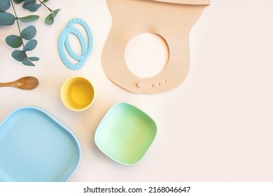 Baby Accessories And Tableware For Food On Beige Background. First Feeding For Kids Concept. Flat Lay, Top View, Space For Text. Silicone Dishware, Bib, Cup On Neutral Backdrop. Serving Baby Food.