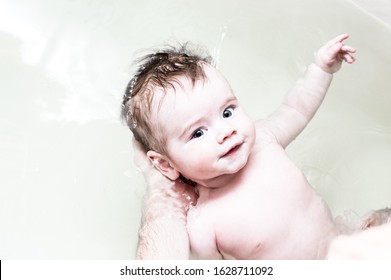 Baby Bathes In The Bath. Dad Holds The Baby In The Water. Infant Smiling