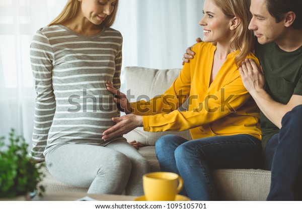 Baby is\
beating. Excited lady is touching abdomen of pregnant woman. Man is\
embracing her and smiling. Surrogacy concept\
