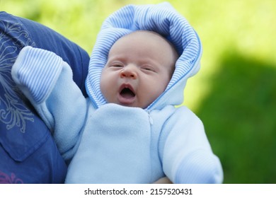A baby in a blue jumpsuit yawns in his mother's arms outside.