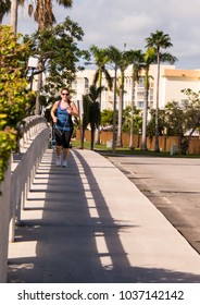 Baby Boomer Woman Power Walking For Exercise Wearing Gym Clothes On A Sidewalk With Palm Trees In Background 