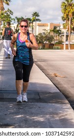 Baby Boomer Woman Power Walking For Exercise Wearing Gym Clothes On A Sidewalk With Palm Trees In Background 