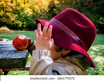 Baby In A Bordeaux Fedora Hat Eating A Red Apple In An Autumn Park Near A Picnic Table. Yellow Foliage And Trees In Background. No Face.