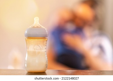 Baby Bottlefeed Concept: Focus On A Bottle With A Mother Holding Her Baby In The Background