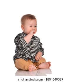 Baby boy in beige pants and a checkered black and white shirt sitting in the studio on a white background. The boy's teeth erupt and he gnaws his fist looking at the free space for text.