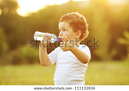 baby boy with brown hair drinking water in the park, holding plastic bottle and showing thumb up. outdoor shot