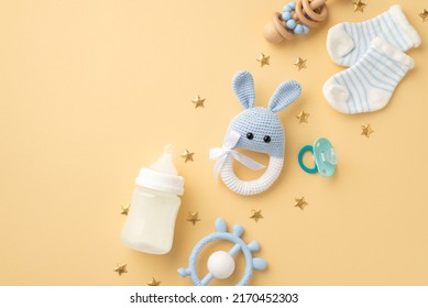 Baby boy concept. Top view photo of knitted bunny rattle toy blue teether wooden rattle milk bottle tiny socks pacifier and gold stars on isolated pastel beige background with copyspace