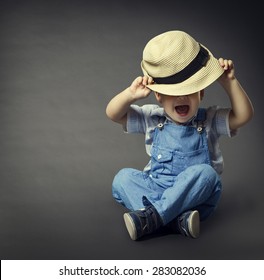 Baby Boy in Fashion Jeans, Hat Covered Eyes. Child Beauty Style, Well Dressed Boy Sitting over Gray Background