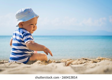 Baby Boy With Hat Sitting On The Beach.