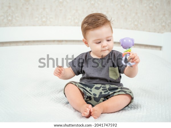 Baby boy with his first toys.\
Charming blond child playing with toy rattle sitting on a\
bed