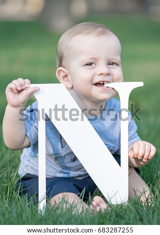 baby boy holding big white letter N and scratching gums on green grass at summer day