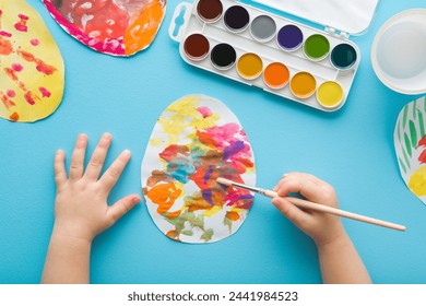 Baby boy holding paintbrush and painting colorful egg on paper with watercolor on light blue table background. Pastel color. Closeup. Child making easter decoration elements. Point of view shot. - Powered by Shutterstock