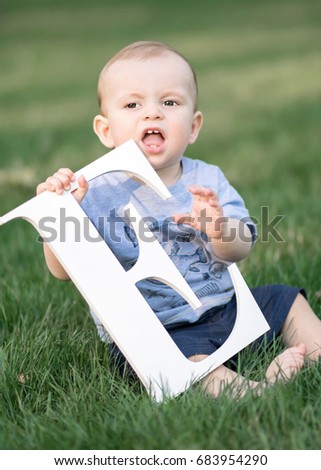 baby boy keeping toy big white letter E sitting on green grass at summer day