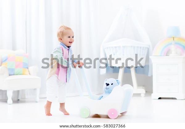 Baby boy learning to walk with wooden push walker in\
white bedroom with pastel rainbow color toys. Aid toy for child\
first steps. Toddler kid walking with car wagon. Nursery interior\
for baby