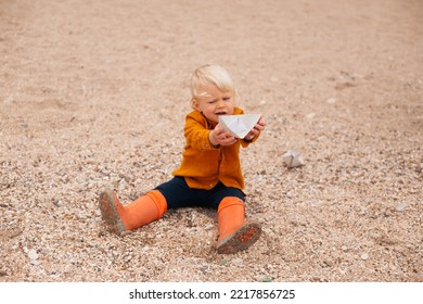 Baby Boy Playing With Paper Boat On The Sandy Beach Near Sea In Autumn Or Summer Time