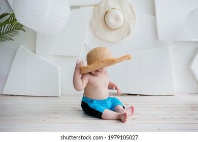 Baby Boy Sits In Blue Shorts And Closed Straw Hat On White Background With Space For Text