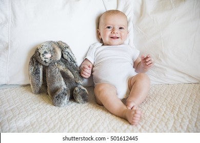 Baby Boy Sits On Bed In A White Onesie With Toy Bunny And Smiles At Camera