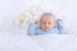 Baby Boy Sleeping On The Bed Lying On His Back With A Stuffed Toy Hares In Blue Pajamas Hands Up, Healthy Newborn Sleep