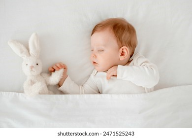 A baby boy sleeps under a blanket in a white bodysuit on the bed with his hand folded under his cheek and holding a soft toy, a sweet healthy baby sleep at home.