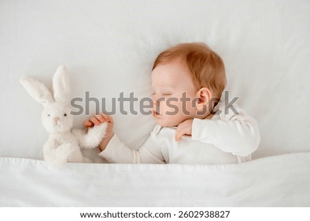A baby boy sleeps under a blanket in a white bodysuit on the bed with his hand folded under his cheek and holding a soft toy, a sweet healthy baby sleep at home.