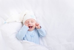 Baby Boy Smiling In His Sleep On The Bed Under The Blanket In A Cap, Healthy Newborn Sleep