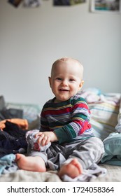 Baby Boy Toddler In A Striped Colour Bodysuit Sitting On A Messy Bed With Clean Clothes Around In The Bedroom And Laughing And Smiling, Happy Expression