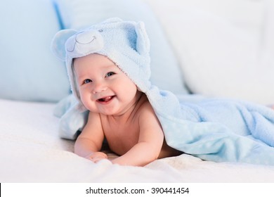 Baby boy wearing diaper and blue towel in white sunny bedroom. Newborn child relaxing in bed after bath or shower. Nursery for children. Textile and bedding for kids. New born kid with toy bear.