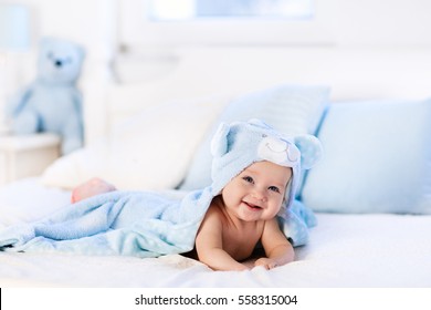 Baby boy wearing diaper and blue towel in white sunny bedroom. Newborn child relaxing in bed after bath or shower. Nursery for children. Textile and bedding for kids. New born kid with toy bear