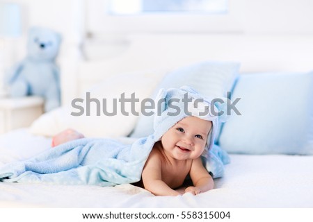 Baby boy wearing diaper and blue towel in white sunny bedroom. Newborn child relaxing in bed after bath or shower. Nursery for children. Textile and bedding for kids. New born kid with toy bear