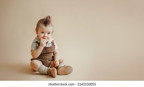 Baby In A Brown Vintage Corduroy Jumpsuit, Shorts, Knee Socks Sits On A Beige Background And Eats Cookies