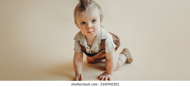 Baby In A Brown Vintage Corduroy Jumpsuit, Shorts, Knee Socks Crawling On The Floor With A Cheerful Grimace And A Cookie In His Hand On A Beige Background