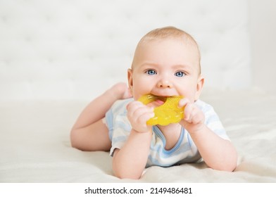 Baby Chewing Teether Toy Lying Down On Stomach In White Bedroom. Child Sucking Nibbler And Massaging Gums. Babies Teething. Cute Six Months Baby Boy Looking At Camera. First Teeth Growth And Care