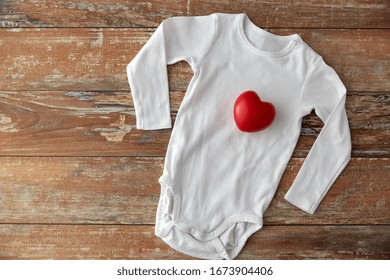 Baby Clothes, Babyhood And Clothing Concept - Bodysuit With Red Heart Toy On Wooden Table