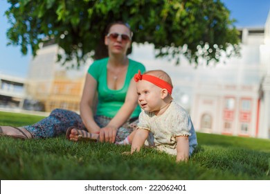 Baby Crawling On The Grass On A Background Of Sitting His Mom. The Kid Runs Away From His Mother. Mother Playing With Her Child.